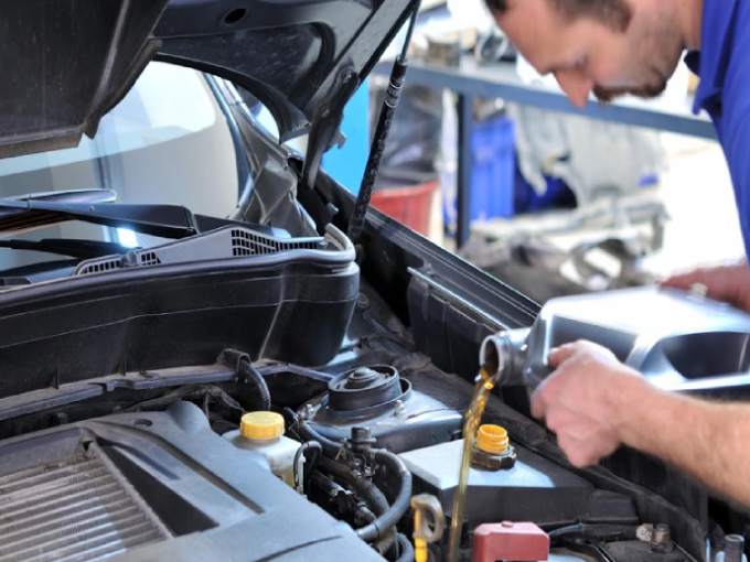 A person pours engine oil into a car’s engine bay under an open hood, performing vehicle maintenance in a workshop setting.